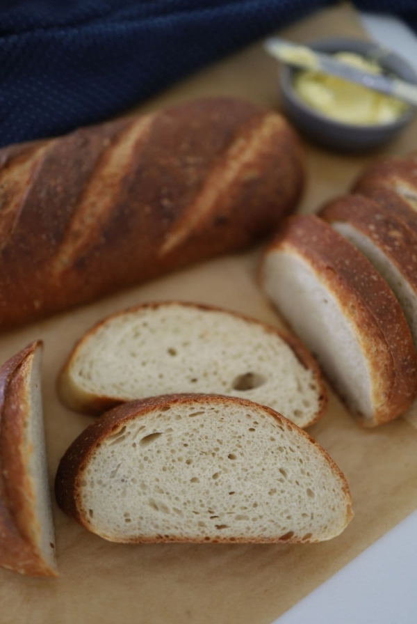 Sourdough French Bread that has been sliced and laid out on a piece of parchment paper. There is a dish of butter in the background of the photo.