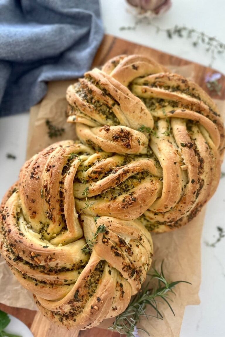 Sourdough Garlic & Herb Twist loaf sitting on a wooden board with some parchment paper. There is a demin blue dish towel in the background.