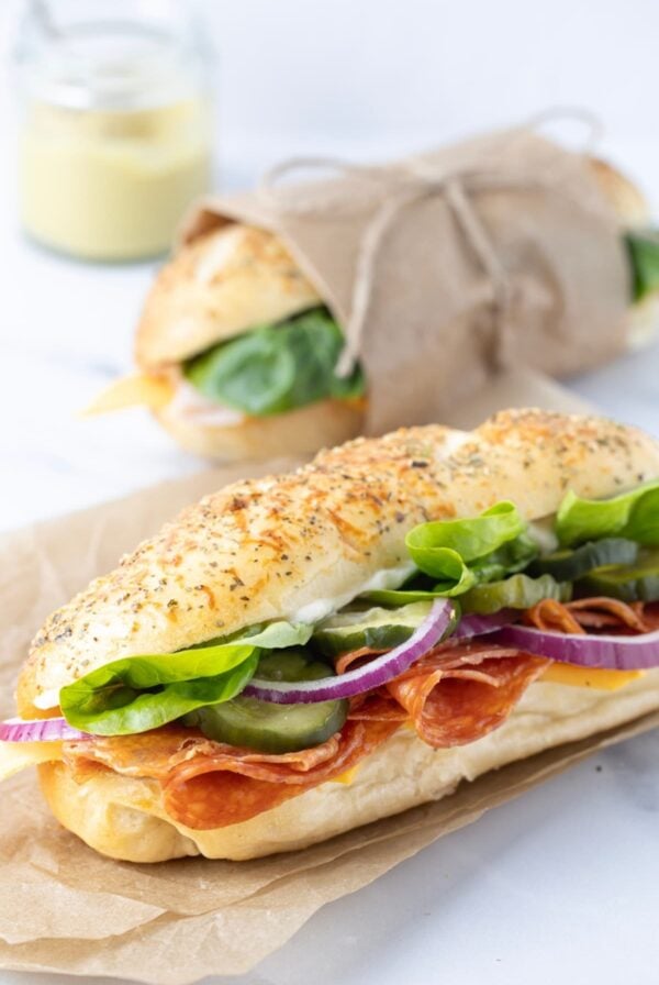 A sourdough sandwich roll filled with salami and salad. The sourdough sandwich roll is topped with Italian herbs. The sandwich roll at the back has been wrapped in parchment paper and tied with jute twine.