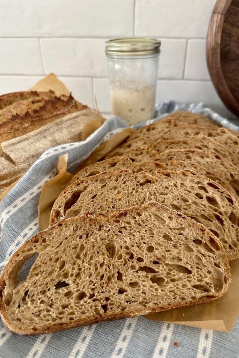 Loaf of sourdough country bread that has been sliced and laid on a blue striped dish towel. There is a jar of sourdough starter in the background.