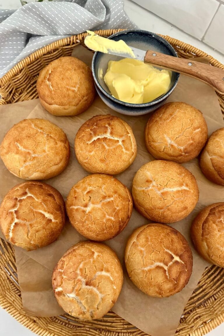 A large cane basket containing 11 sourdough tiger rolls all laid out so you can see the crackly crust. There is a dish of butter and a small butter spreader in the basket also.