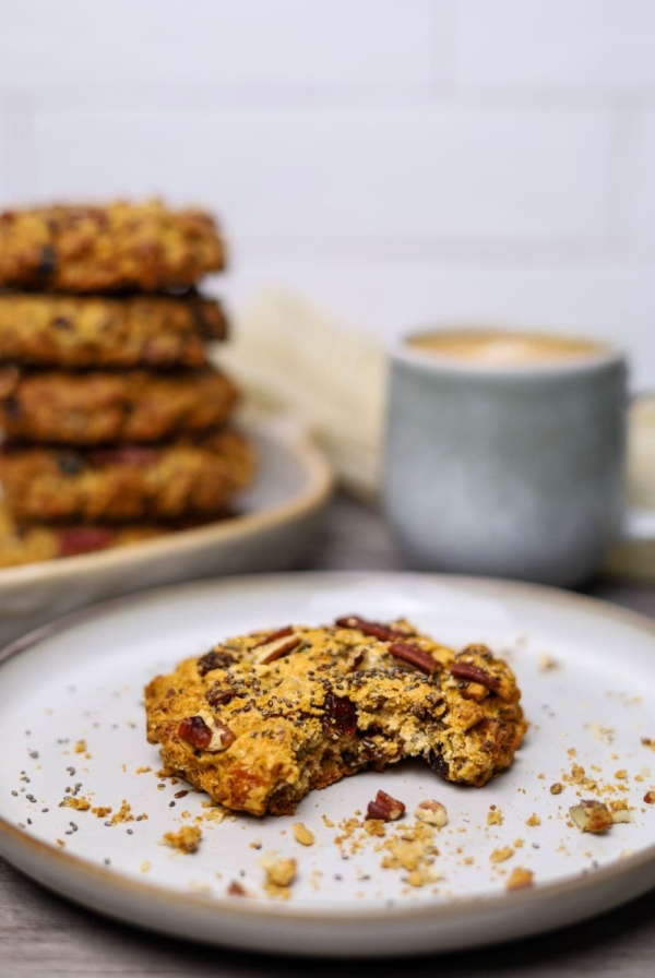 A sourdough breakfast cookie on a beige plate. There is a bite taken out of the cookie and it is surrounded by crumbs. In the background you can see a stack of 5 other cookies and a mug of coffee.