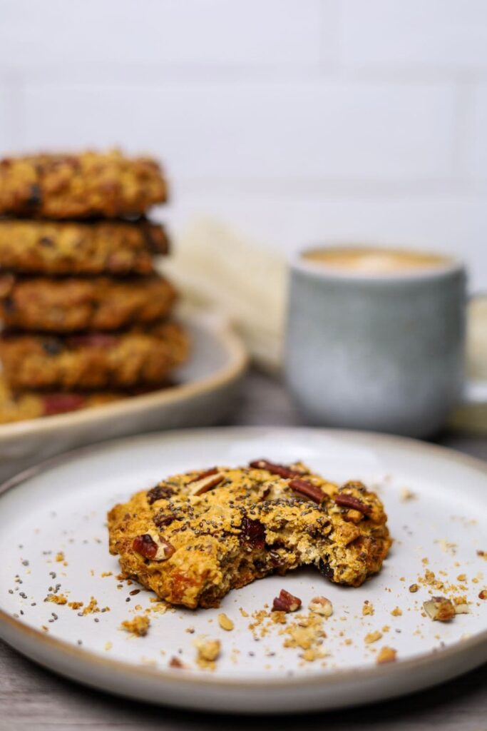 A sourdough breakfast cookie on a beige plate. There is a bite taken out of the cookie and it is surrounded by crumbs. In the background you can see a stack of 5 other cookies and a mug of coffee.