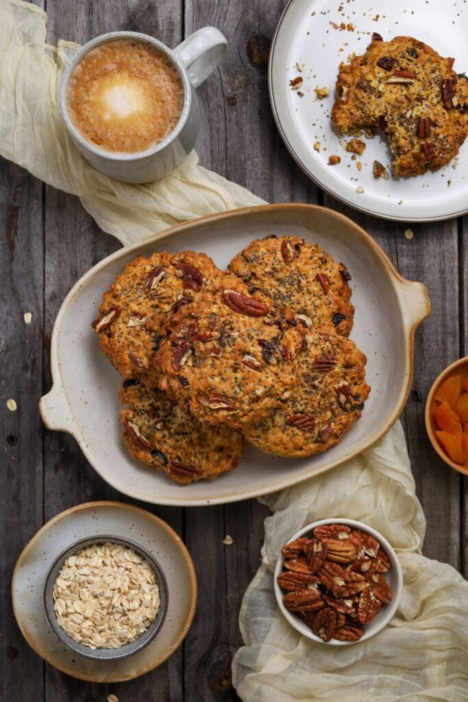 Flat lay of a rustic breakfast scene featuring a platter of sourdough breakfast cookies, surrounded by a cup of coffee, a bowl of dried apricots, a bowl of pecans, and a bowl of rolled oats on a dark wooden surface. A plate containing a partially eaten cookie and a soft cream-colored cloth add texture and warmth to the setup.