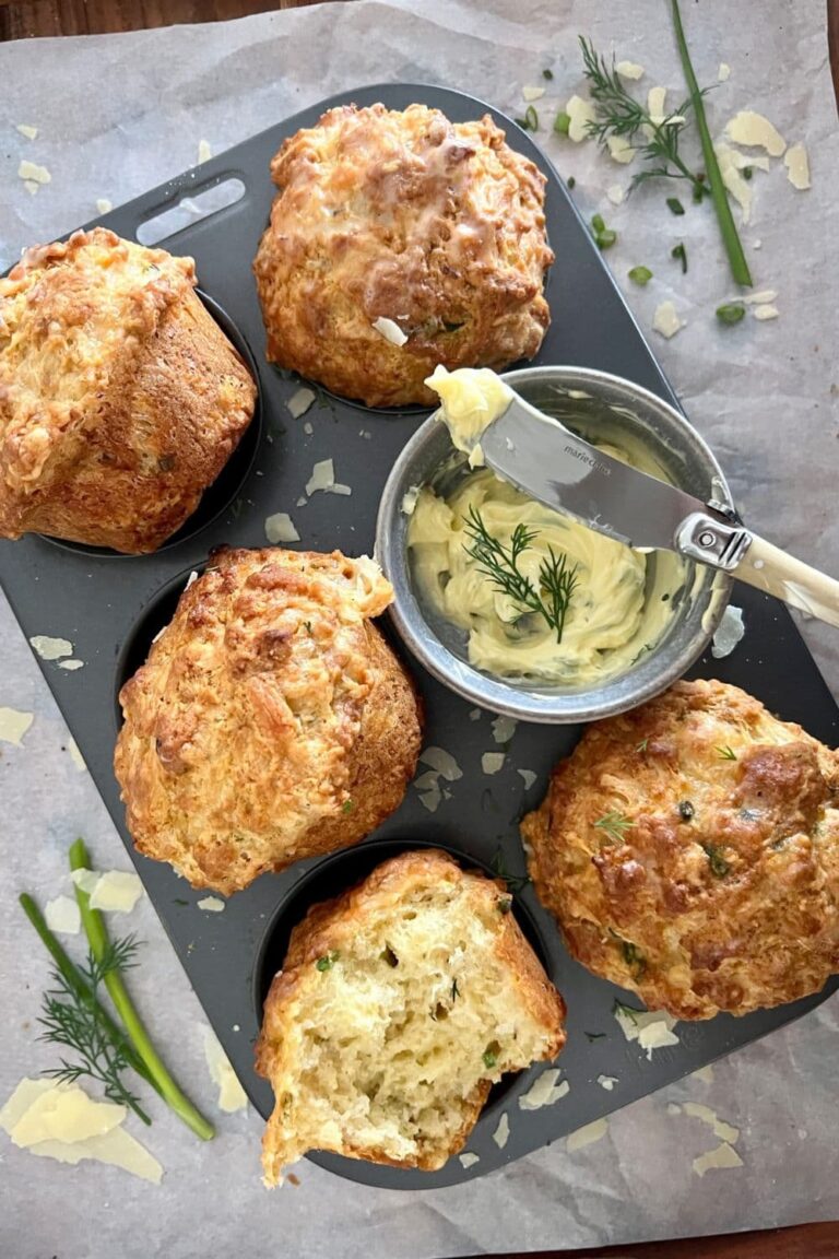 Sourdough Cheese Muffins sitting in a muffin tin. There is a bowl of herbed butter sitting in the muffin tin. There is dill and parmesan cheese sprinkled around the muffins.