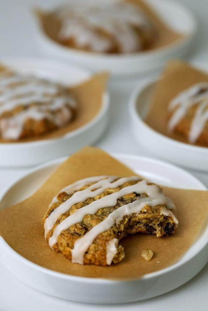 A sourdough oatmeal raisin cookie drizzled in a simple sugar glaze sitting on a piece of parchment paper on a small white plate.