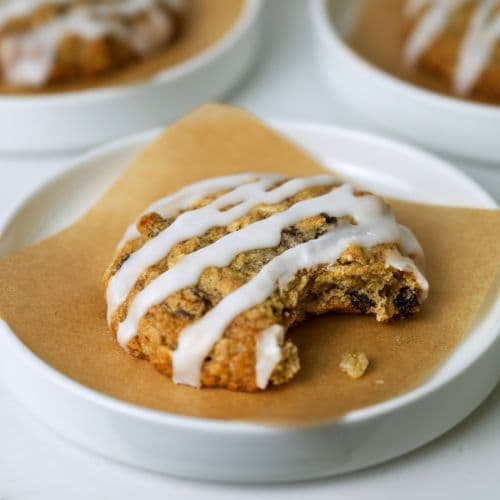 A sourdough oatmeal raisin cookie drizzled in a simple sugar glaze sitting on a piece of parchment paper on a small white plate.