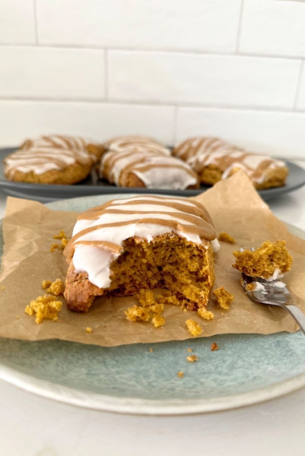 Sourdough pumpkin scone with vanilla glaze and pumpkin spice glaze sitting on a blue stoneware plate. There are a whole plateful of sourdough pumpkin scones in the background.