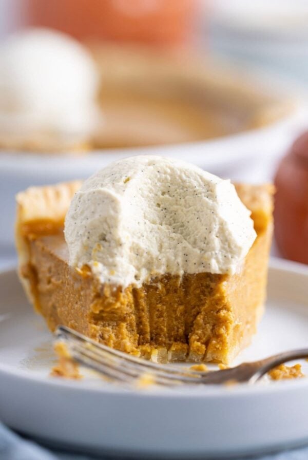 A close up image of a slice of sourdough pumpkin pie topped with vanilla maple whipped cream. There is a piece that has been taken out of the pie with a fork so you can see the silky texture of the pumpkin custard made with sourdough starter.