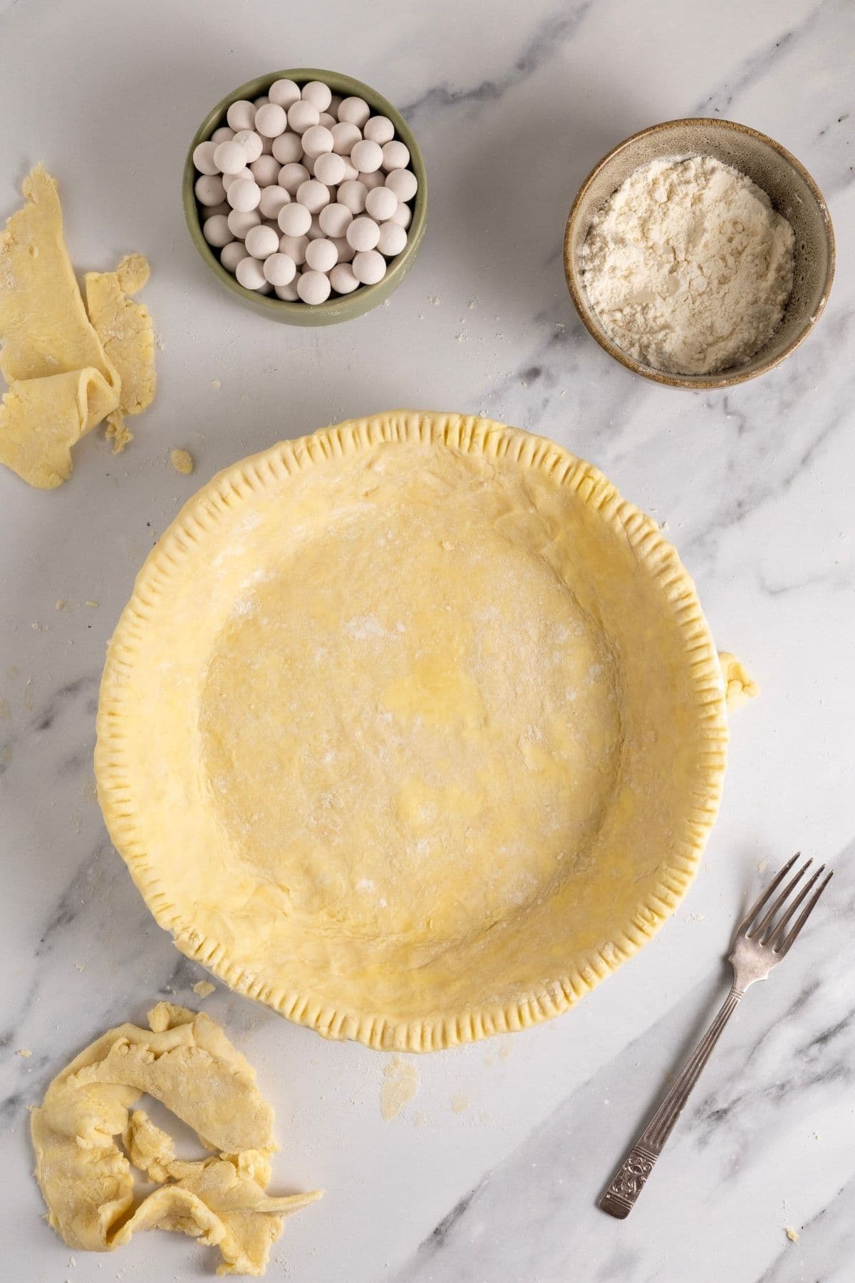 An image showing that the pastry has been trimmed around the edges of the pie plate and the edges then crimped with a vintage fork that is now sitting to the right of the pie dish.
