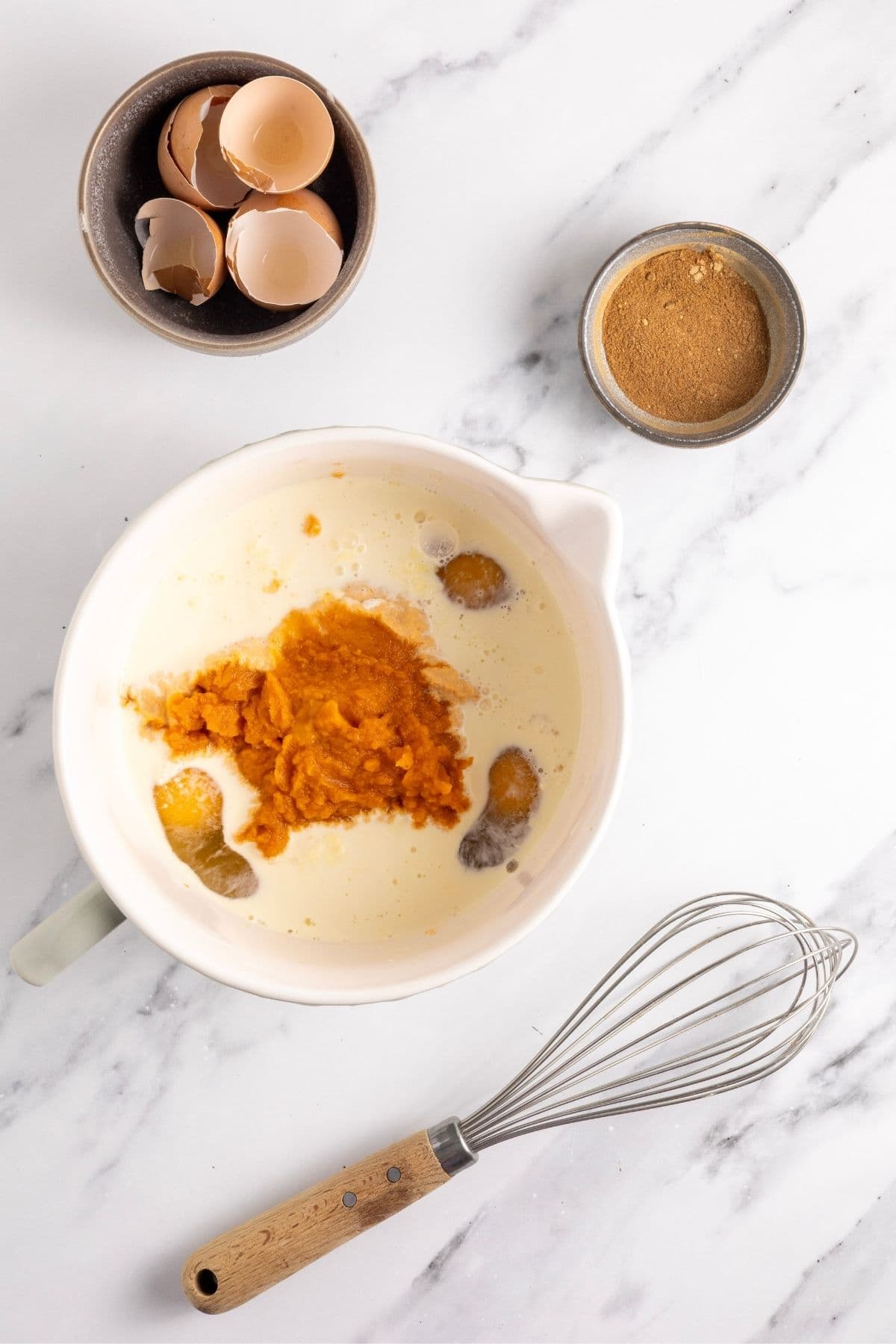 An image showing the sourdough pumpkin pie filling being whisked together. There is a bowl of egg shells and a small bowl of pumpkin pie spice in the top of the image.