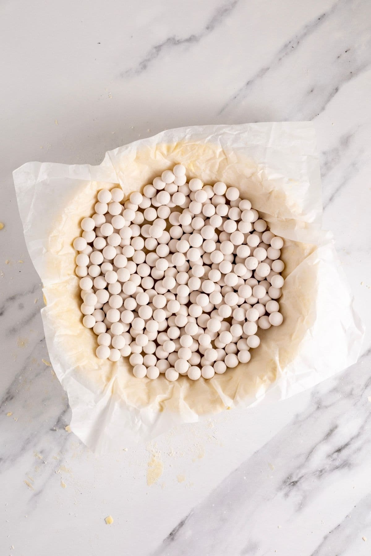 The pie dish filled with pastry that has a piece of white parchment paper filling the cavity that is then filled with white porcelain pie weights. 