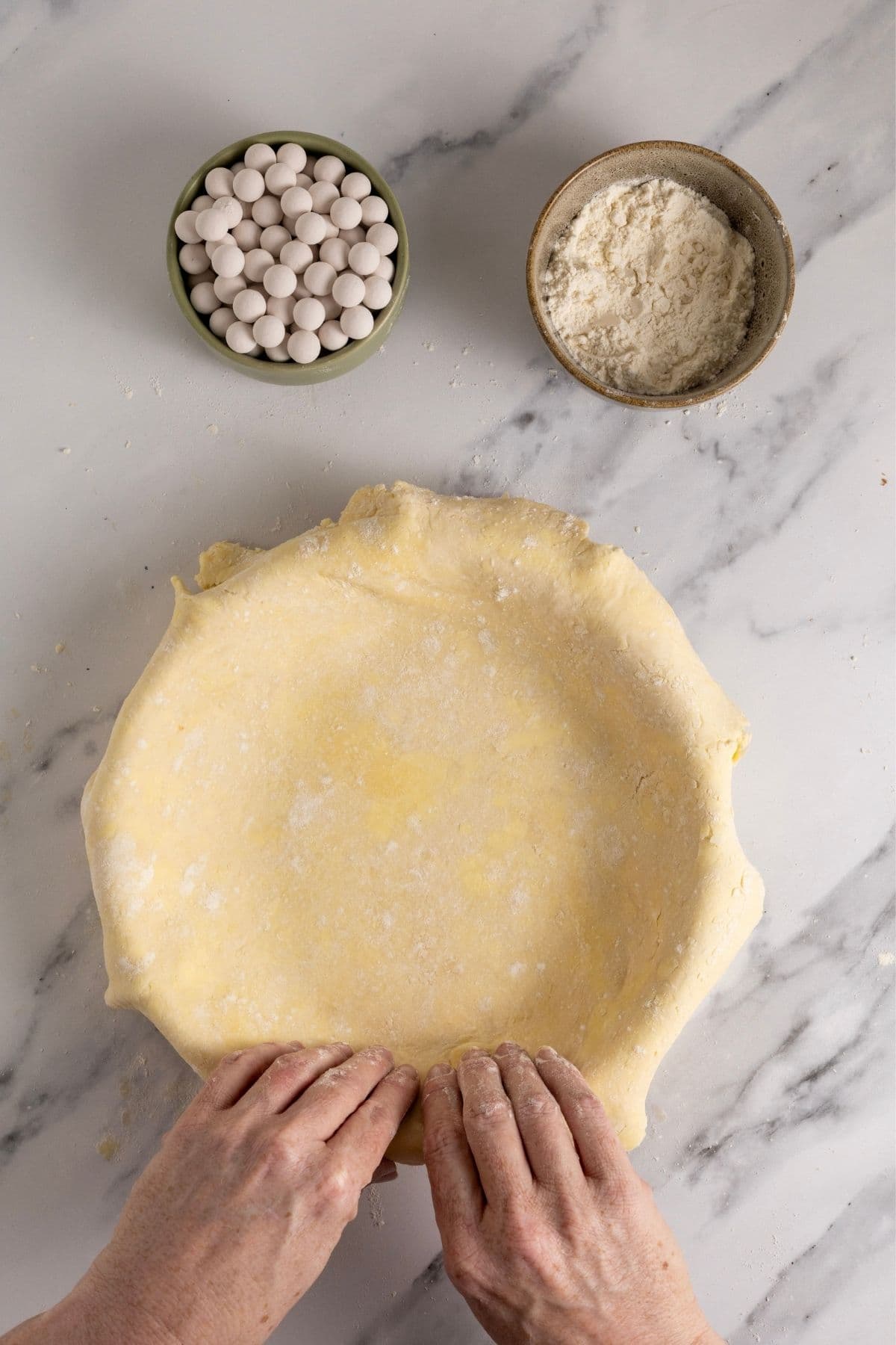 A pie dish that has been filled with sourdough pastry. A pair of hands are pushing the pastry into the base of the pie dish so that it doesn't shrink when it bakes.