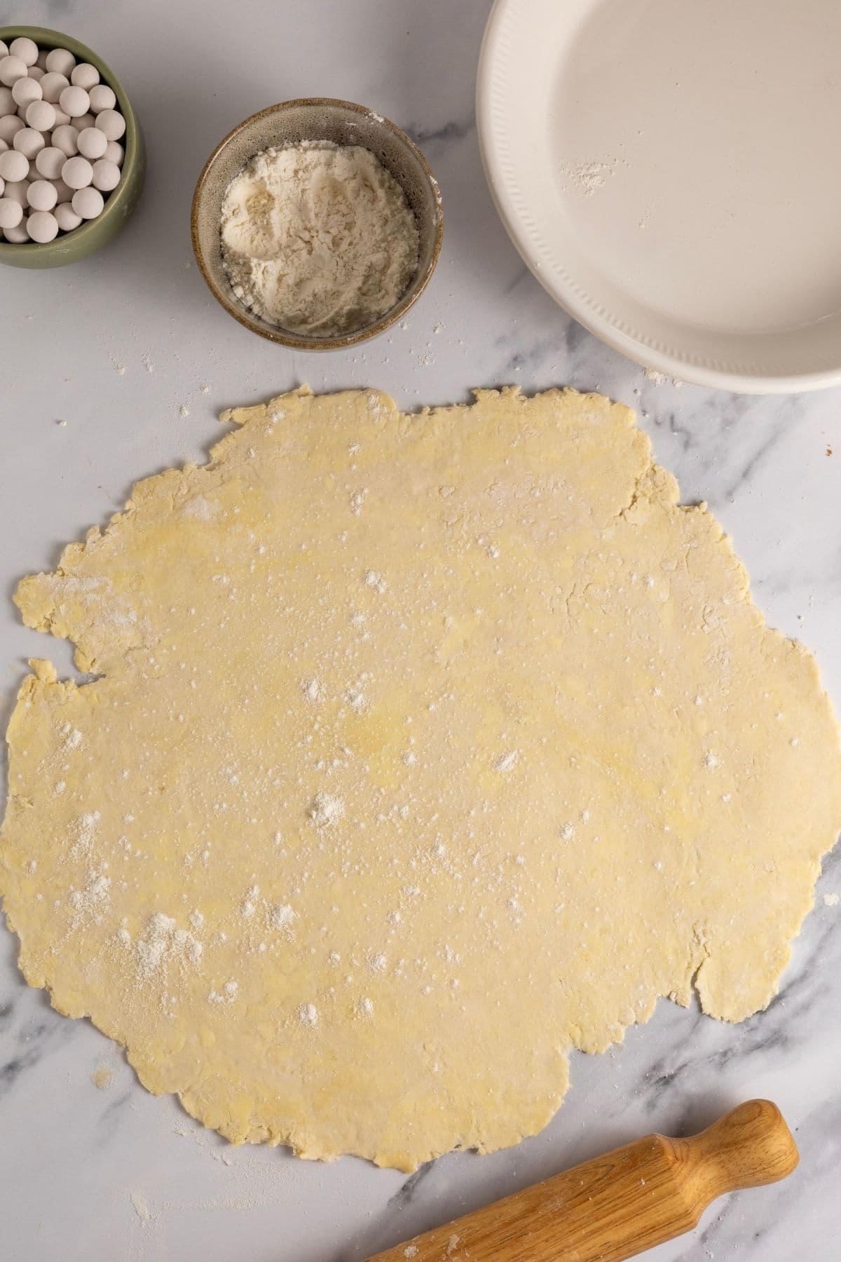 A sheet of sourdough pastry rolled out on the counter top. There is also a small bowl of flour and pie weights in the image.