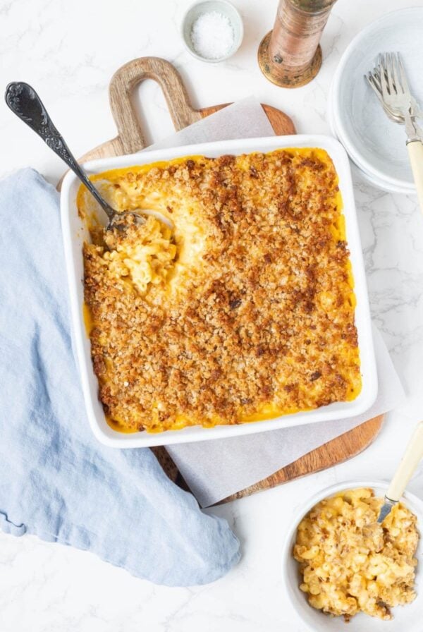 A vertical image showing a square baking dish of sourdough mac and cheese with a spoon digging into the velvety smooth creamy cheese sauce. The dish is topped with crunchy baked sourdough bread crumbs.