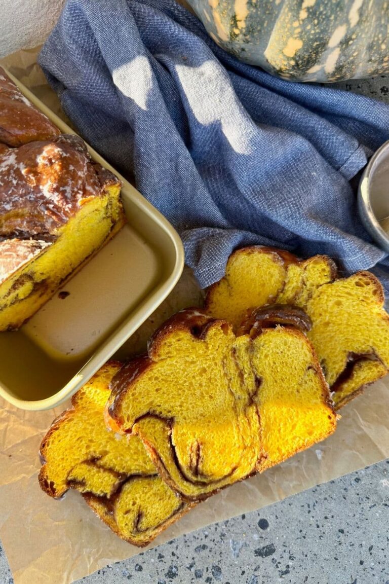 Sourdough pumpkin babka that has been sliced and laid out on a piece of parchment paper. You can see the remainder of the loaf sitting in a gold loaf pan. There is a blue dish towel and a whole pumpkin in the left hand side of the photo.