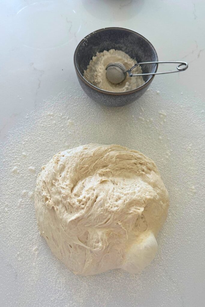 A ball of sourdough ciabatta dough tipped out on the counter with a big bubble. There is a small grey bowl of flour on the counter with a sifter to help with shaping the dough.