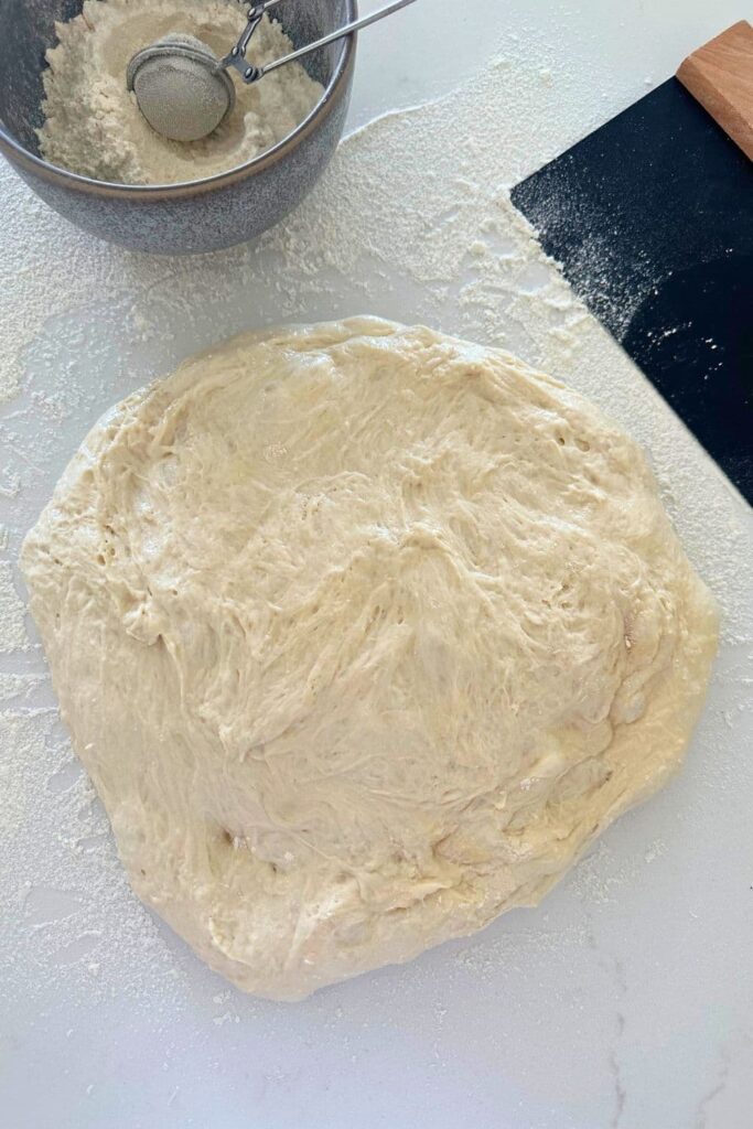 Sourdough ciabatta dough spread out on the counter. There is a small bowl of flour and dough scraper also visible in the photo.