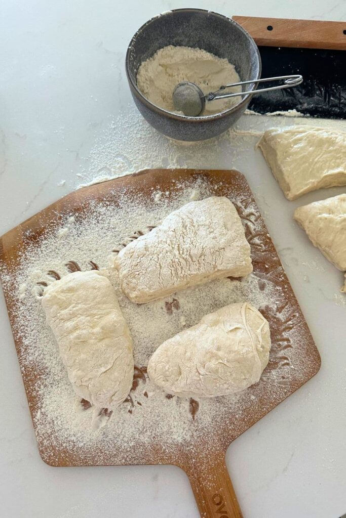 sourdough cibatta dough sitting on a heavily floured pizza peel. You can see a bowl of flour with a tea strainer sitting in the photo, along with a black steel dough scraper.