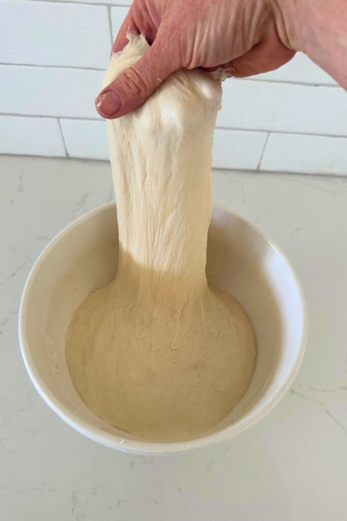 A bowl of sourdough ciabatta dough that is being stretched out of the bowl.