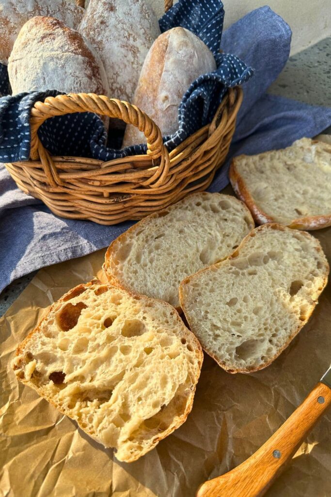 A basket of sourdough cibatta rolls. There is a cibatta roll that has been sliced in half sitting in front of the basket.