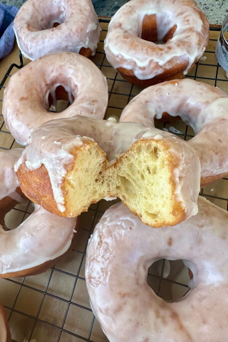 Fried sourdough donuts with vanilla glaze laid out on a wooden board. One of the donuts has a bite taken out so you can see the light, yeasty inside of the donut.