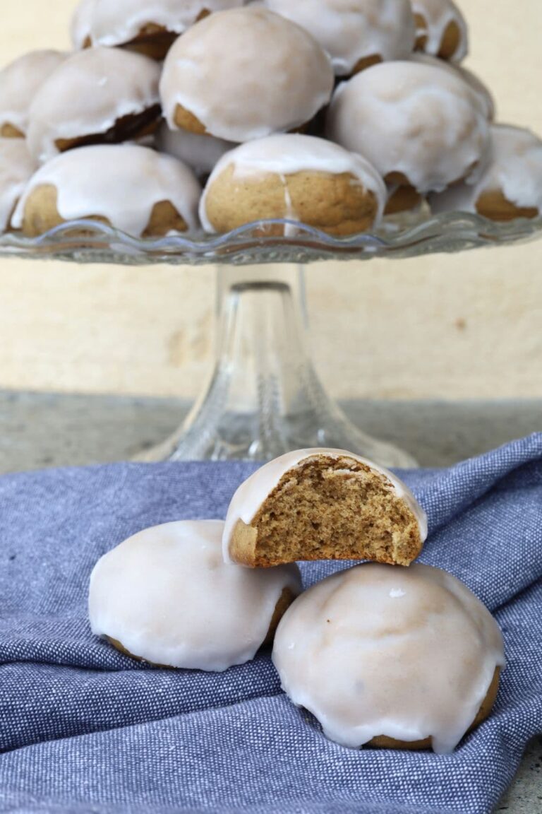 Sourdough pfeffernusse cookies sitting on a glass cake plate. There is a blue dish towel underneath with a few pfeffernusse sitting on them.