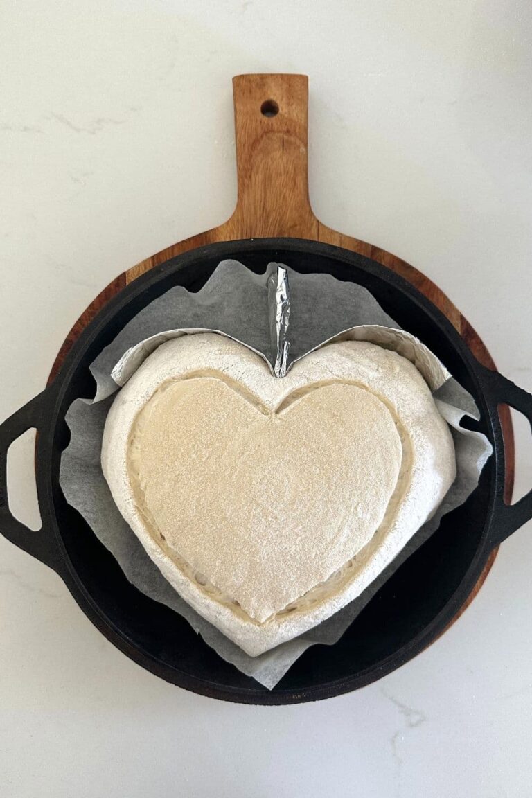 Heart shaped sourdough bread sitting in a Lodge Cast Iron Dutch Oven skillet lid. The dough has been scored with a large heart and has some aluminium foil in the Dutch Oven to help the bread heart retain its shape.