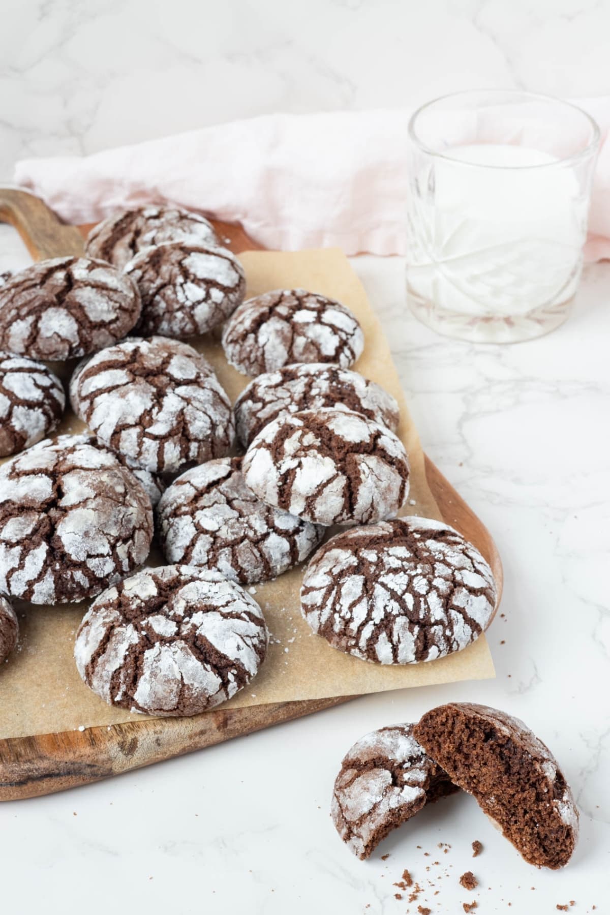 A vertical image showing a pile of sourdough chocolate crinkle cookies displayed on a wooden board. There is a glass of milk at the side of the board and a cookie broken in half to show the texture in side at the front of the photo.