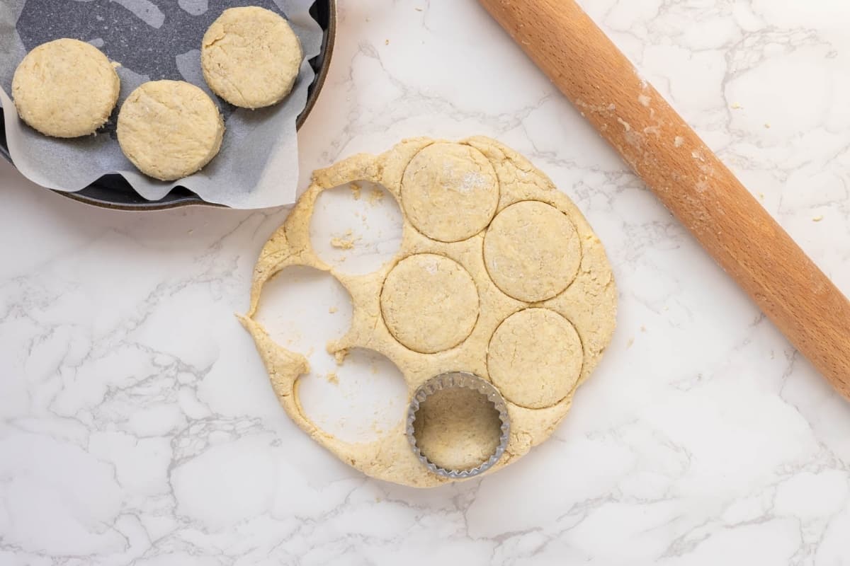 Showing the biscuit dough being cut into rounds using a biscuit cutter. You can see the bottom half of a cast-iron skillet with three of the dough rounds sitting in it.