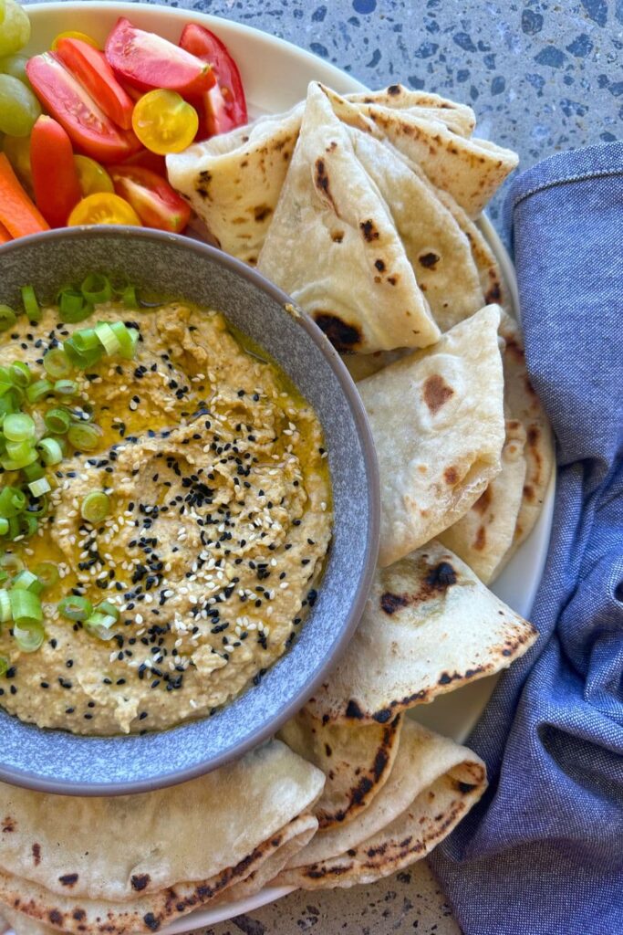 A bowl of hummus made without tahini. It's served in the middle of a platter surrounded by sourdough flat bread, red and yellow tomatoes and is topped with chopped shallots.