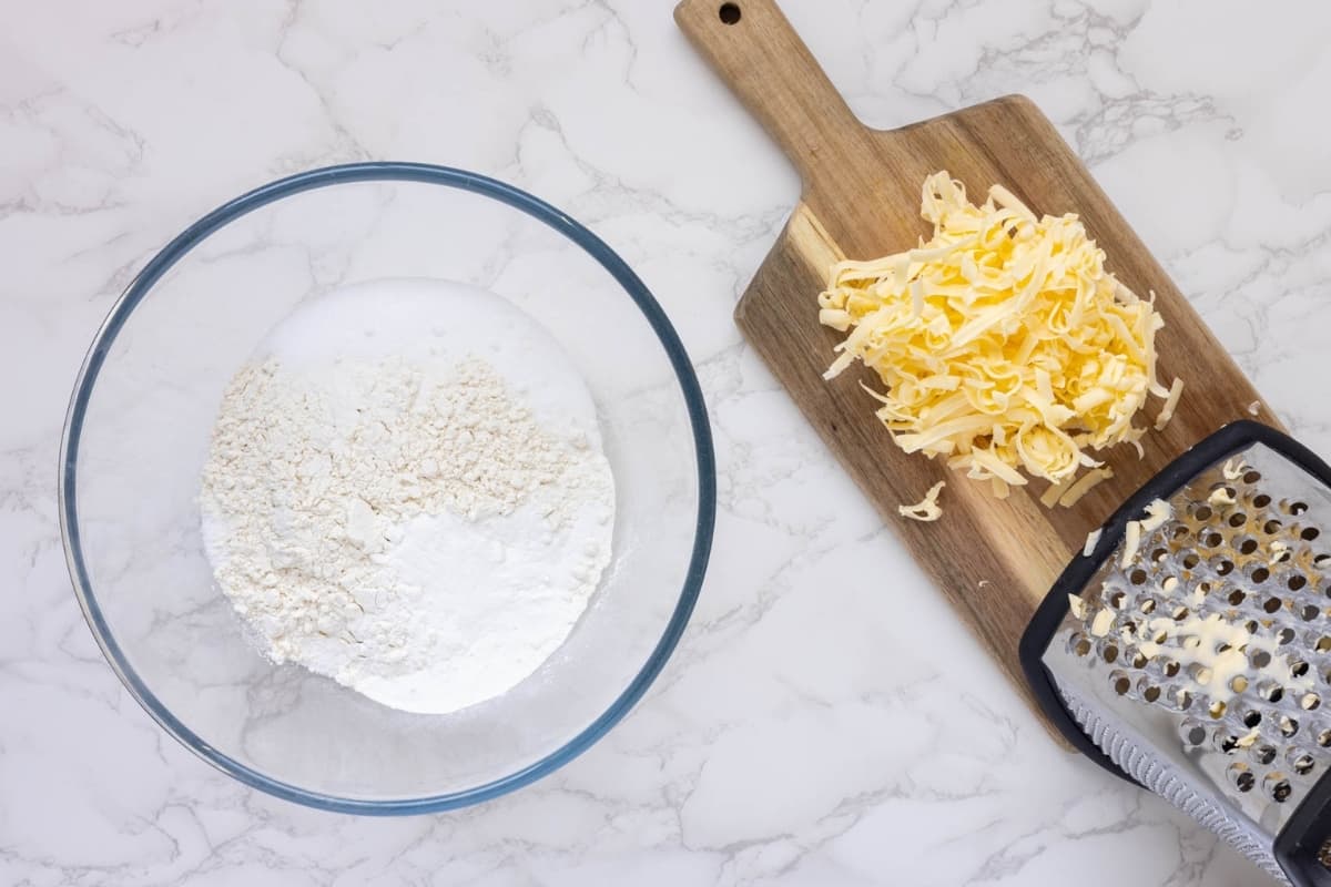 A glass bowl filled with the dry ingredients sitting next to a pile of yellow butter that has been grated on a box grater.