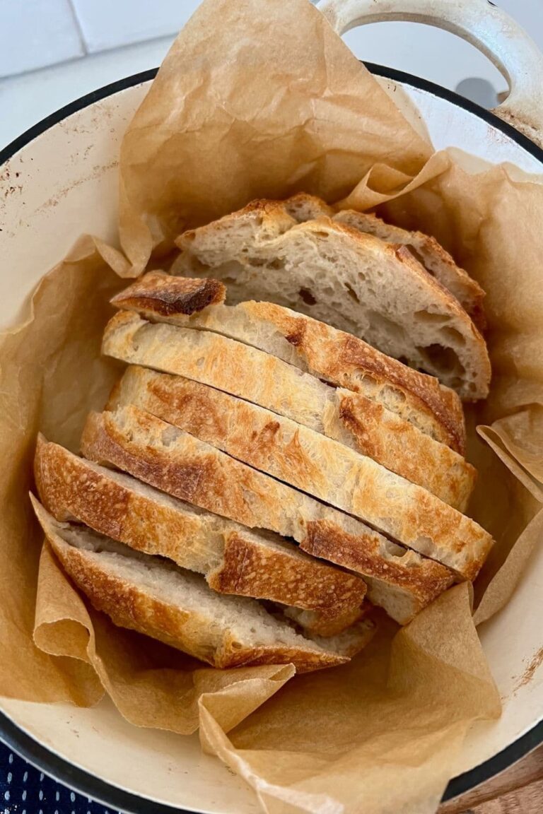 A loaf of sourdough bread that has been slice and is nestled in a piece of parchment paper inside a cream enamel Dutch Oven.