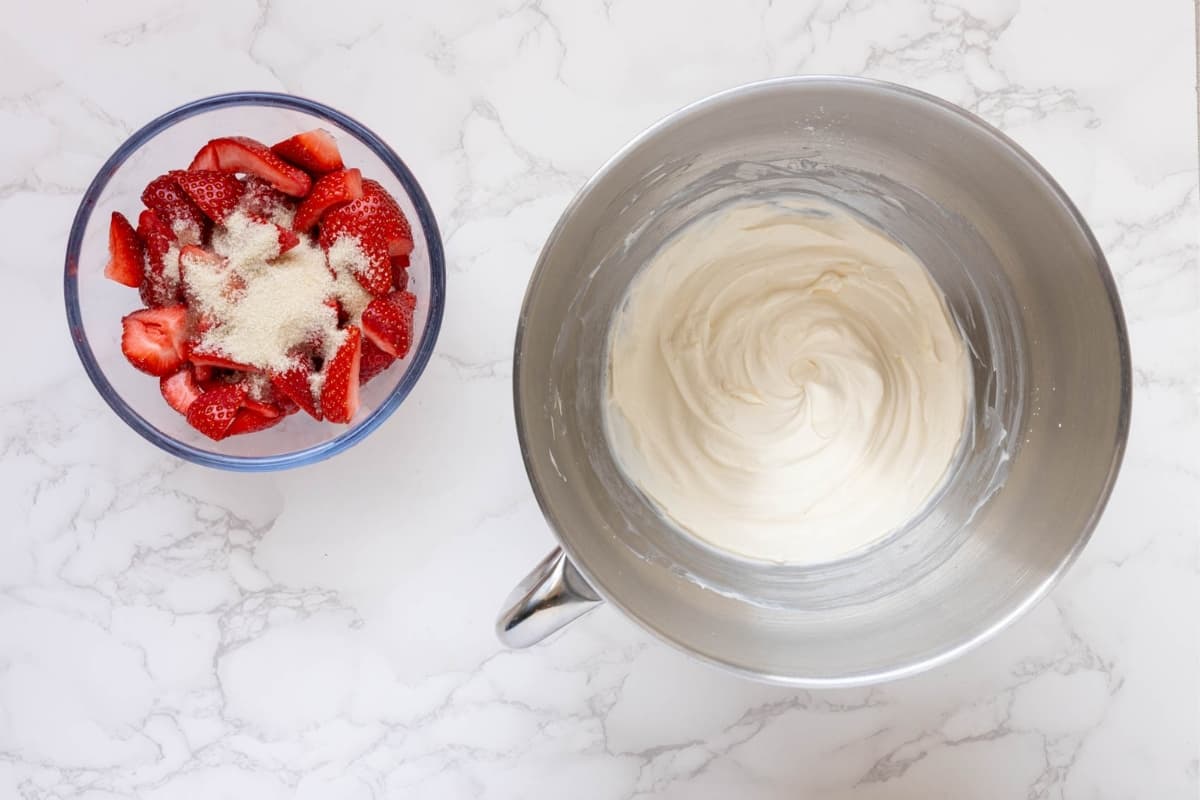 A photo showing the sliced strawberries sitting in a bowl with the sugar. There is a stainless steel stand mixer bowl sitting to the right filled with vanilla whipped cream. 