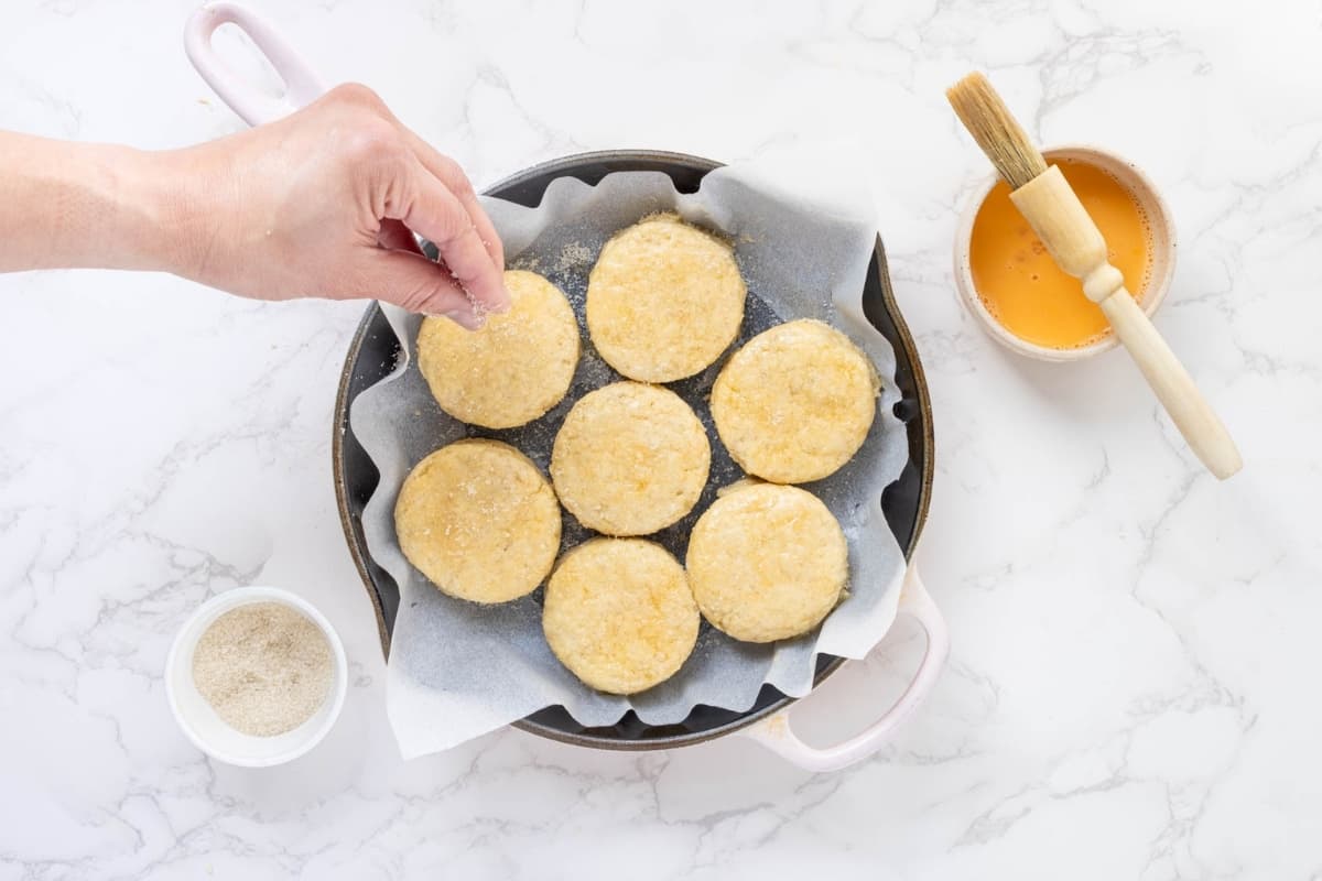 An image showing a cast-iron skillet lined with parchment paper and filled with 7 sourdough biscuits. There is a hand sprinkling sugar on top. To the right, there is a small bowl of egg wash with a pastry brush sitting on top,.