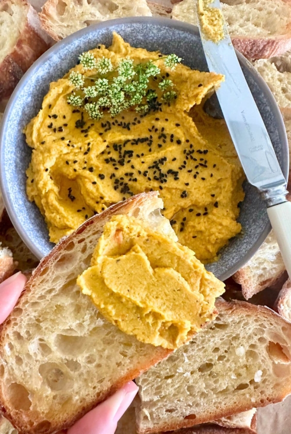 A bowl of roasted vegetable hummus decorated with cumin seeds. The bowl is surrounded by sourdough crostini and there is a slice of sourdough crostini spread with roasted carrot hummus in the foreground.
