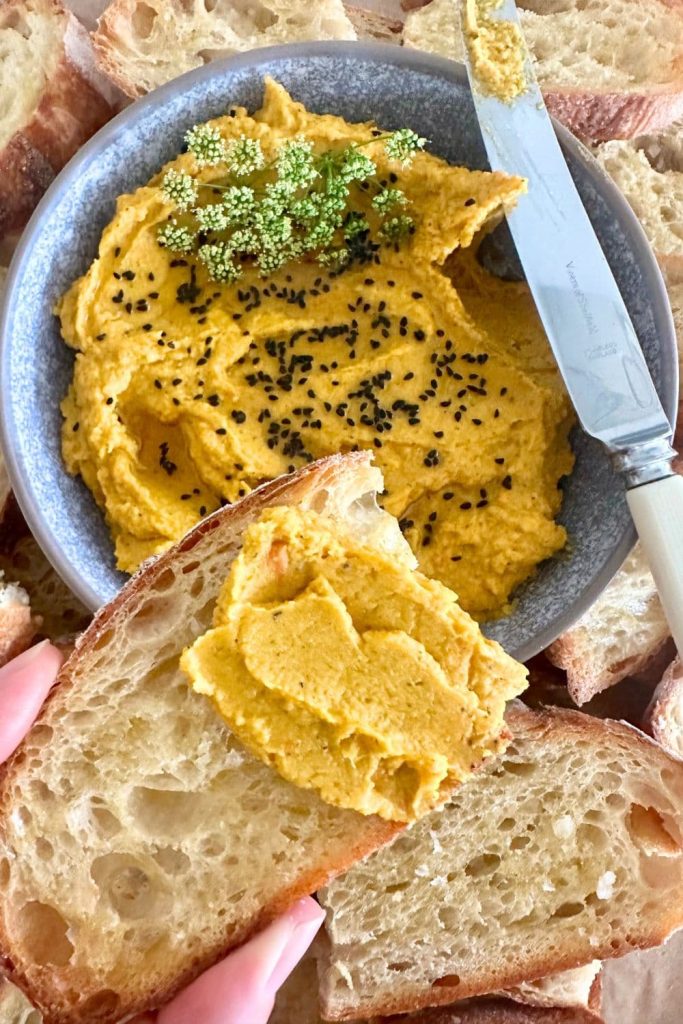 A bowl of roasted vegetable hummus decorated with cumin seeds. The bowl is surrounded by sourdough crostini and there is a slice of sourdough crostini spread with roasted carrot hummus in the foreground.