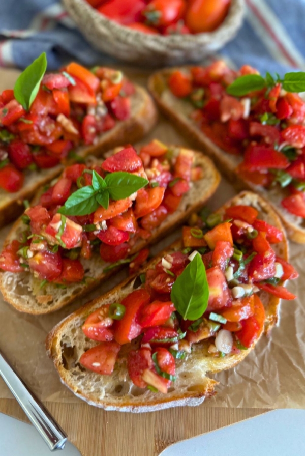 4 slices of sourdough bruschetta sitting on a piece of parchment paper. There is a basket of Roma tomatoes sitting to the left of the photo.