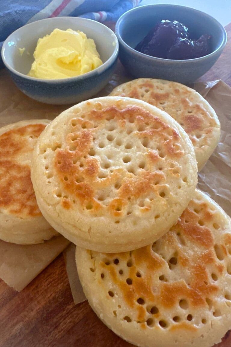 4 sourdough crumpets displayed on a wooden board. There are 2 bowls of butter and jam sitting behind the sourdough crumpets.