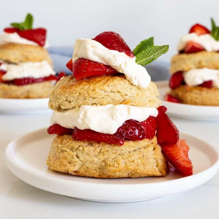 A close up image of a strawberry shortcake made with sourdough biscuits, whipped cream and juicy red strawberries. It is garnished with fresh, green mint leaves.