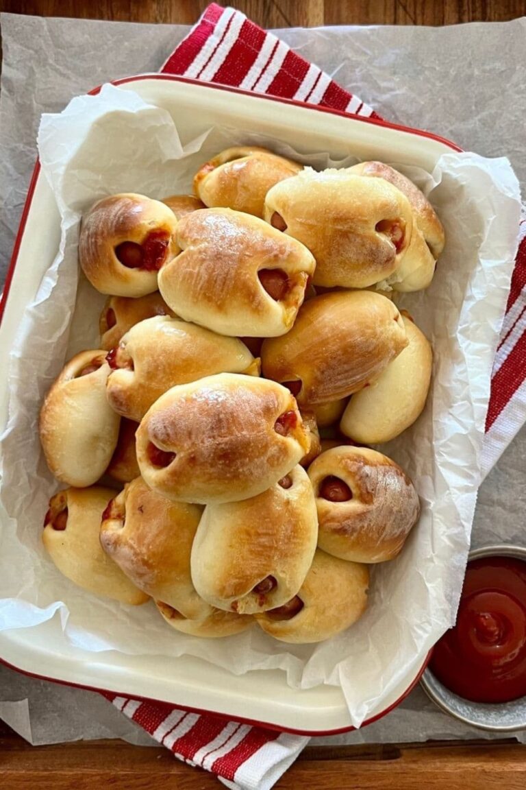 A dish of sourdough pigs in blankets ready for sourdough super bowl snacking. The cream enamel dish is sitting on a red striped dish towel. There is a bowl of tomato ketchup to the right of the sourdough pigs in blankets.