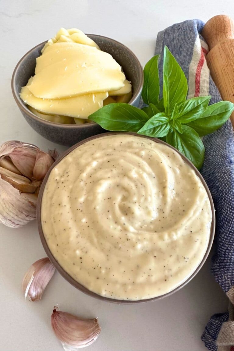 A bowl of white pizza sauce made with sourdough starter sitting on a white counter top. The bowl is surrounded by garlic cloves, cheese and basil leaves.