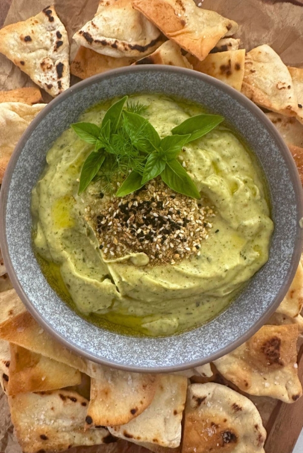 Zucchini yogurt dip topped with dukkah, olive oil and fresh herbs and served in a grey stoneware bowl. The bowl of zucchini yogurt dip is surrounded by sourdough pita chips.