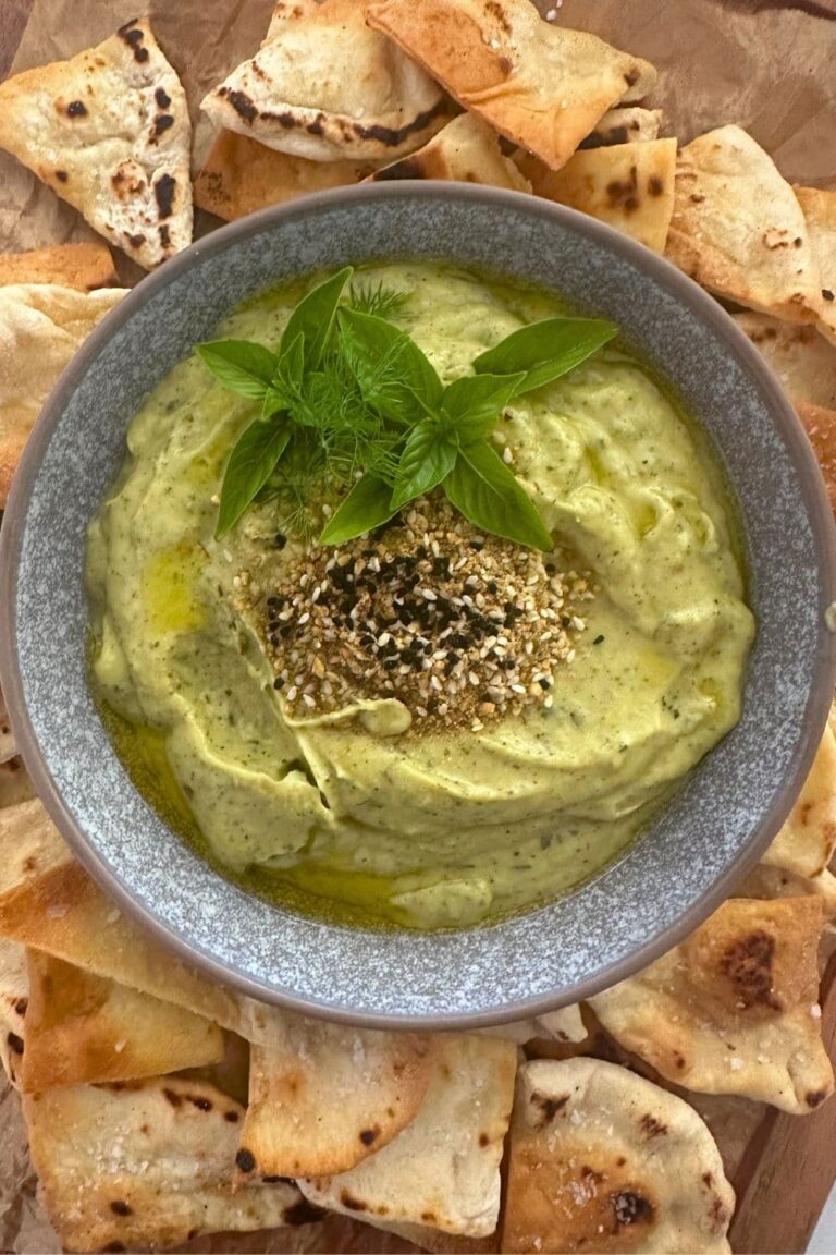 Zucchini yogurt dip topped with dukkah, olive oil and fresh herbs and served in a grey stoneware bowl. The bowl of zucchini yogurt dip is surrounded by sourdough pita chips.