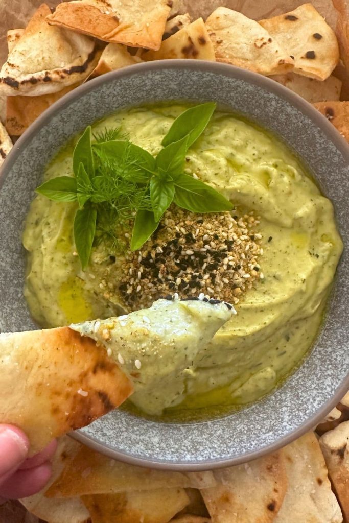 A bowl of bright green zucchini yogurt dip topped with cumin seeds, dukkah and some fresh herb leaves. It's being dipped into by a sourdough pita chip.