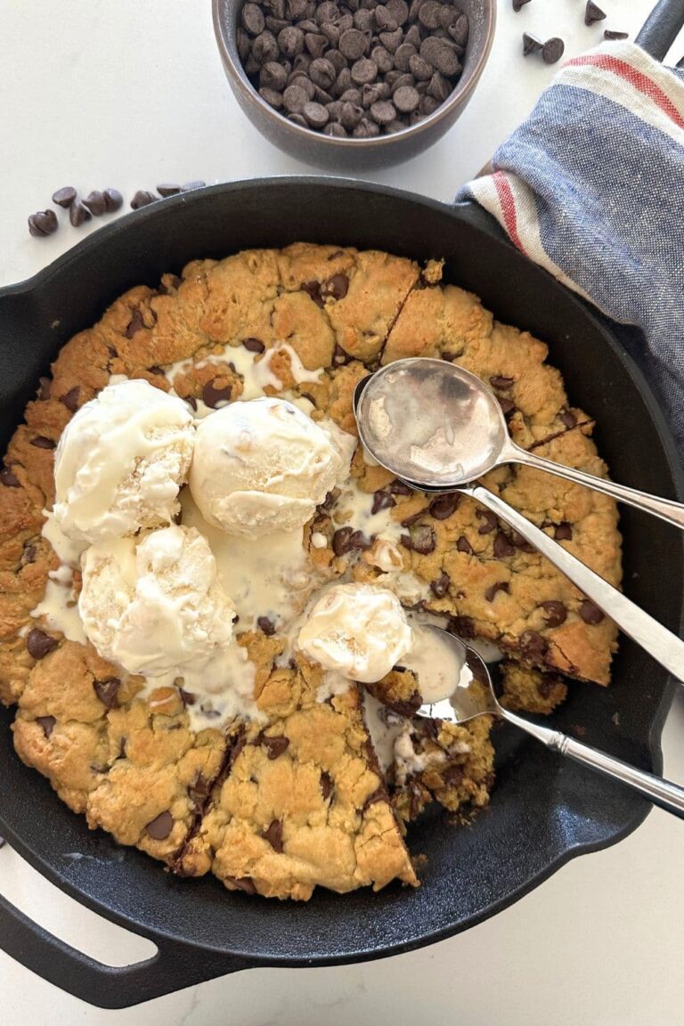 Sourdough Cookie Skillet with chocolate chips - Recipe Feature Image. This image is of a cast iron skillet filled with a giant sourdough chocolate chip cookie baked in the oven and then topped with vanilla ice cream. There are three spoons in the skillet and some of the giant cookie has been eaten.