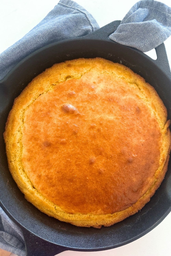 A cast-iron skillet containing sourdough cornbread that has just come out of the oven. It is golden brown and surrounded by a blue dish towel.