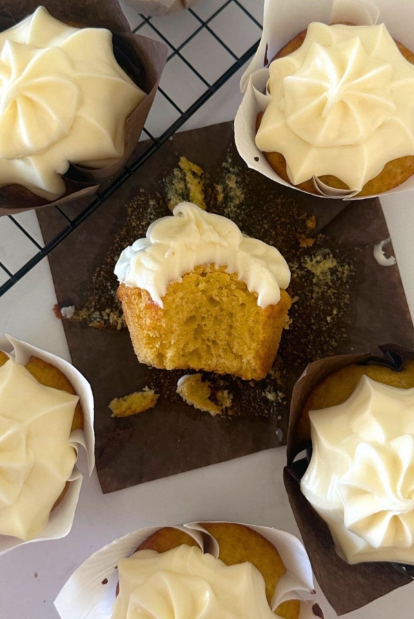 Sourdough vanilla cupcakes displayed on a black wire cooling rack. The cupcake in the middle has been bitten into so you can see the moist, buttery crumb. All of the cupcakes have been frosted with vanilla cream cheese frosting.