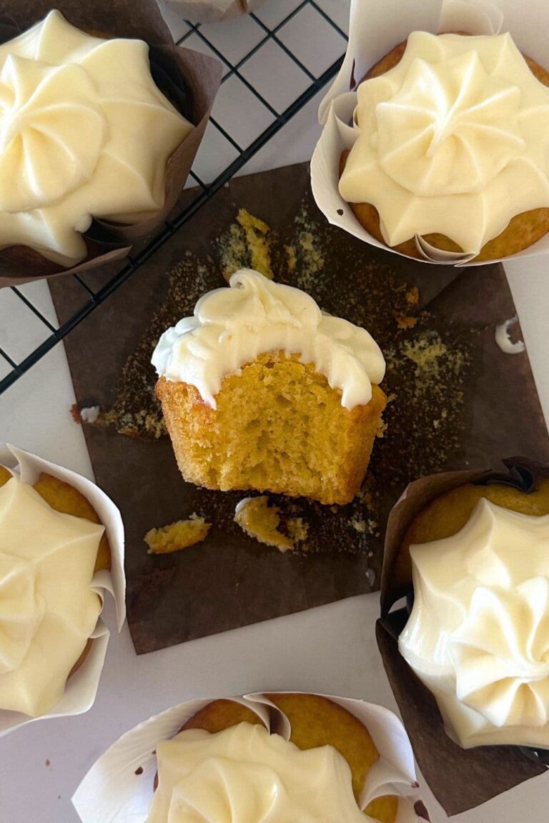 Sourdough vanilla cupcakes displayed on a black wire cooling rack. The cupcake in the middle has been bitten into so you can see the moist, buttery crumb. All of the cupcakes have been frosted with vanilla cream cheese frosting.