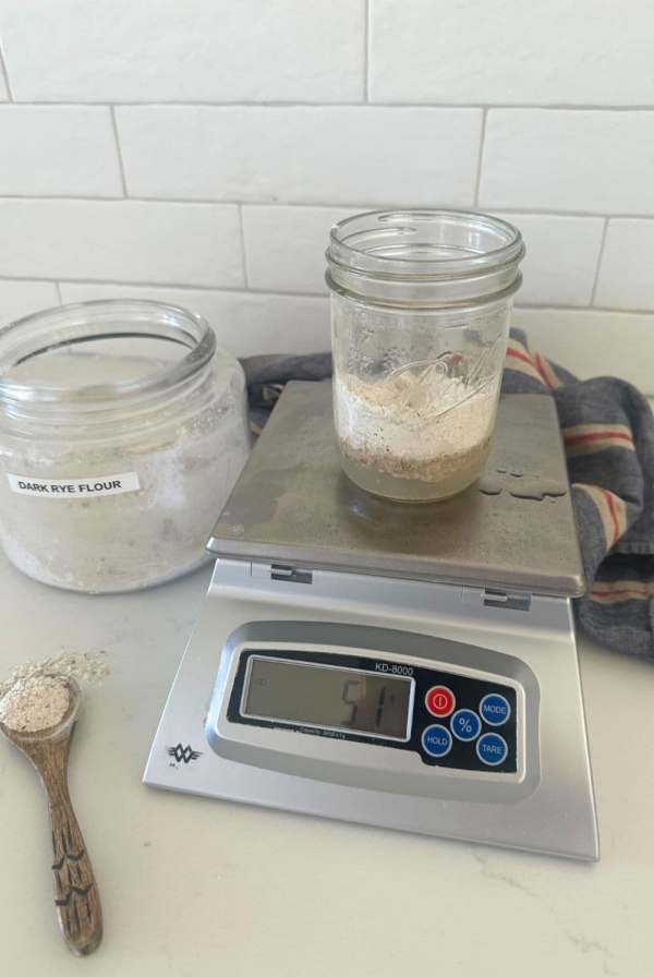 A photo of a 16oz mason jar sitting on a scale. The jar is filled with water and dark rye flour to create a rye flour sourdough starter. You can also see a large jar of rye flour in the background and a wooden spoon with rye flour on it next to the scale.